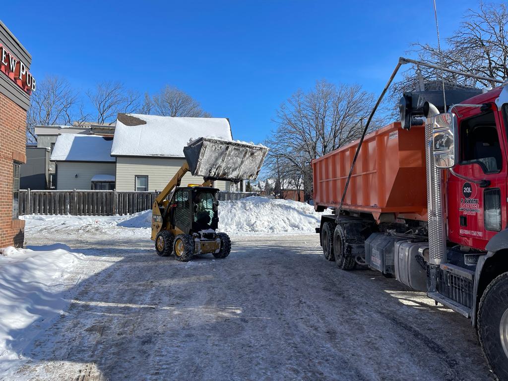 A skid steer loader is moving snow onto a waiting dump truck, showcasing a strategic snow clearance operation in a residential area, keeping the streets safe and passable during winter in Ottawa.